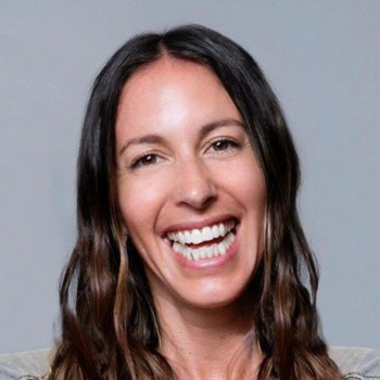 A woman smiling broadly against a plain grey background with long wavy brown hair.